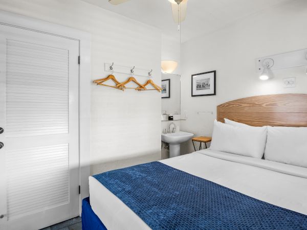 A small hotel room with a bed, blue blanket, two framed pictures, hooks with hangers, and a bathroom sink visible in the corner.