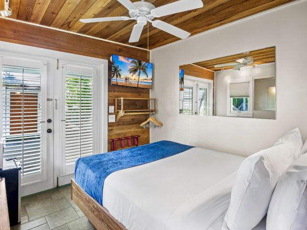 The image shows a cozy bedroom with a double bed, blue blanket, wooden accents, ceiling fan, TV, and a glass door with shutters leading outside.