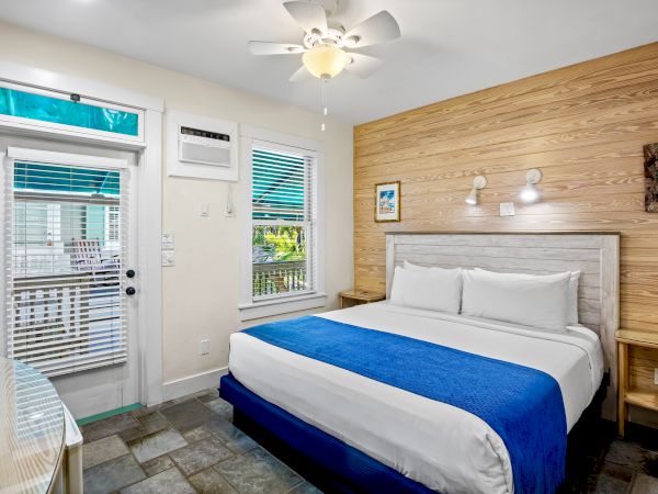 A cozy bedroom with a large bed, blue blanket, two nightstands, window, door, and ceiling fan, featuring wood accent wall and tile floor.