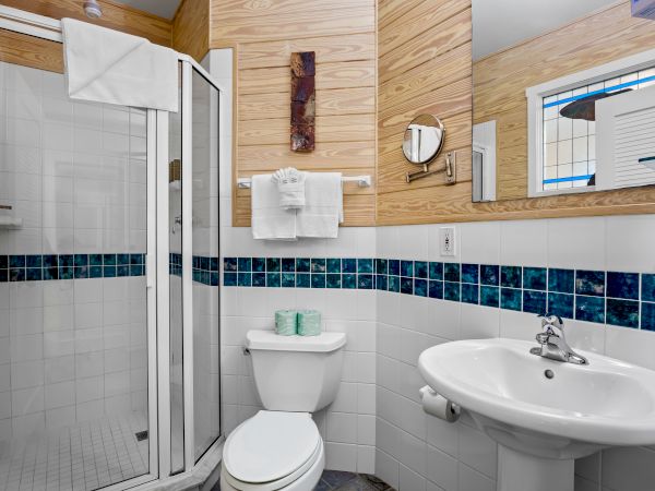 The image shows a small bathroom with a shower, sink, mirror, and toilet. Decorative tile accents and wooden panels are on the walls.