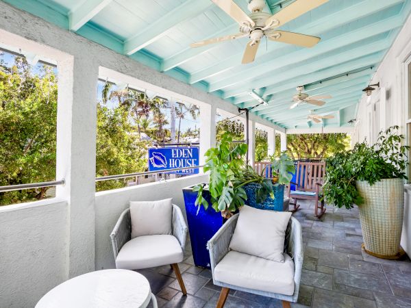 A porch with plants, comfy chairs, a ceiling fan, and a "Eden House" sign, surrounded by greenery and a relaxing atmosphere.