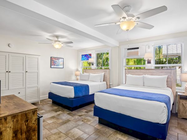 A bright room with two beds, blue accents, ceiling fans, and ample natural light from windows. A TV and furniture are also visible.
