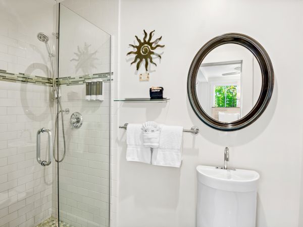 A bathroom with glass shower, round mirror, small sink, towels, and decorative wall piece.