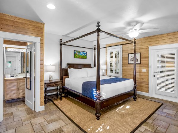 A cozy bedroom with a four-poster bed, blue bedding, wooden walls, a ceiling fan, and a doorway leading to a bathroom.
