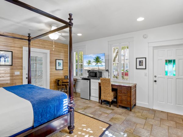 A cozy bedroom with a four-poster bed, desk, chair, microwave, and a TV displaying a beach scene. Light wood walls and tile flooring complete it.