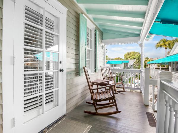 The image shows a porch with two wooden rocking chairs, white railings, French doors, and teal accents. It's a bright, relaxing setting.