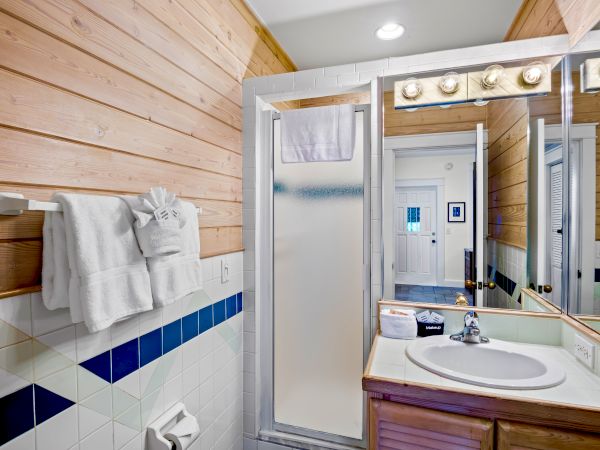 A bathroom with wood-paneled walls, a shower, a sink, and a mirror. Towels hang next to the shower along with a wall-mounted light fixture.