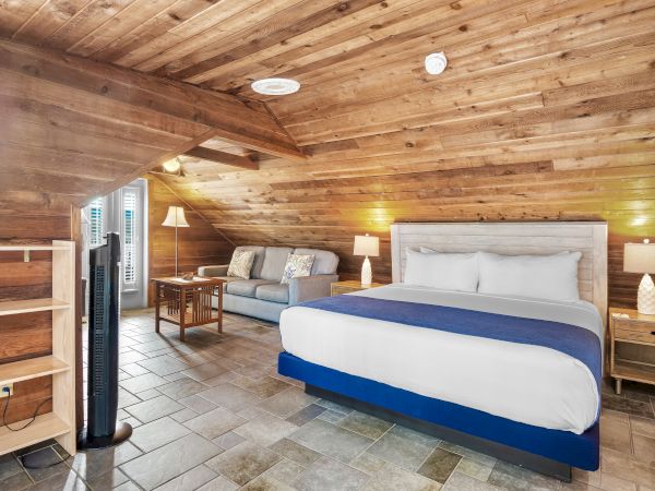 The image shows a cozy attic room with a bed, a couch, wooden walls and ceiling, a stand fan, and two bedside lamps for lighting.