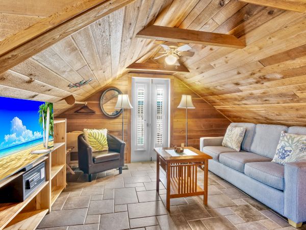 A cozy attic room with wooden walls, a TV, sofa, armchair, coffee table, and a ceiling fan, featuring a bright window.