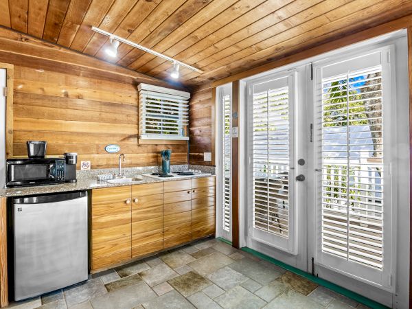 A cozy kitchen with wooden walls features a small fridge, sink, coffee maker, and microwave, leading to a patio through French doors.