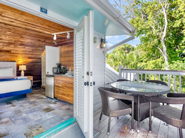 A cozy room with wood accents opens to a balcony featuring a round table and chairs, surrounded by lush greenery in the background.