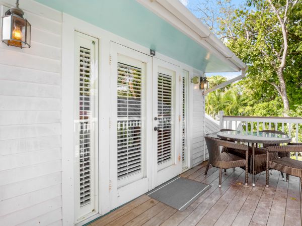 A wooden deck with a small glass table and chairs is alongside double doors with blinds, surrounded by greenery and a wall lantern.