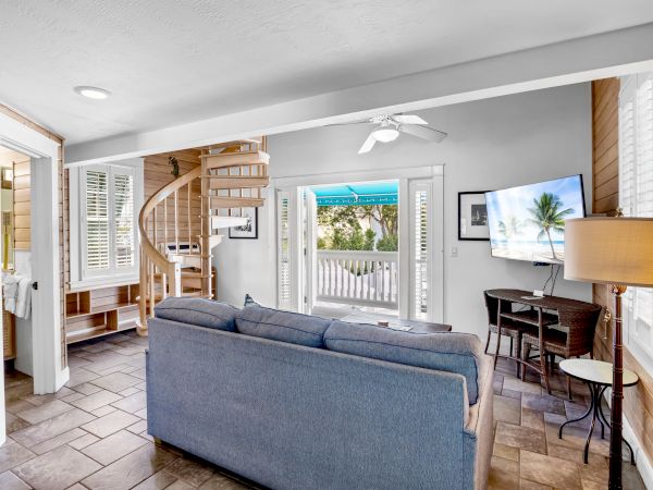 A cozy living room with a spiral staircase, sofa, TV, and bright decor. French doors lead to a porch with palm tree view outside.