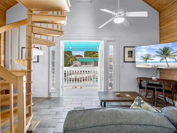 The image shows a cozy living room with a spiral staircase, a ceiling fan, TV, and a view of a balcony with palm trees outside.