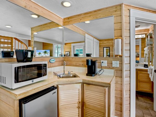 A small kitchenette with a microwave, mini-fridge, coffee maker, sink, and wooden cabinets next to a mirrored wall and a door.