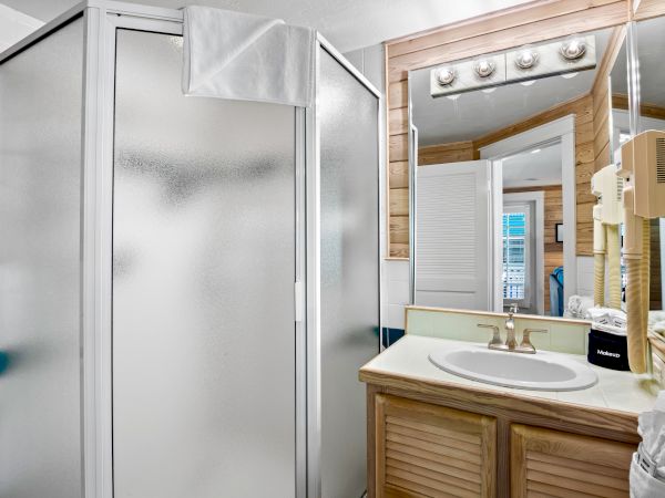 The image shows a bathroom with a frosted glass shower, sink with a mirror, lights above, and wooden cabinets below.
