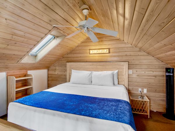 A cozy attic bedroom with wooden ceiling, double bed, blue blanket, ceiling fan, skylight, small shelf, and side table with a fan.