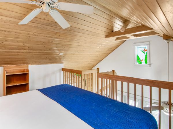 The image shows a cozy loft bedroom with a wooden ceiling, white walls, a bed with a blue blanket, a ceiling fan, and a small window.