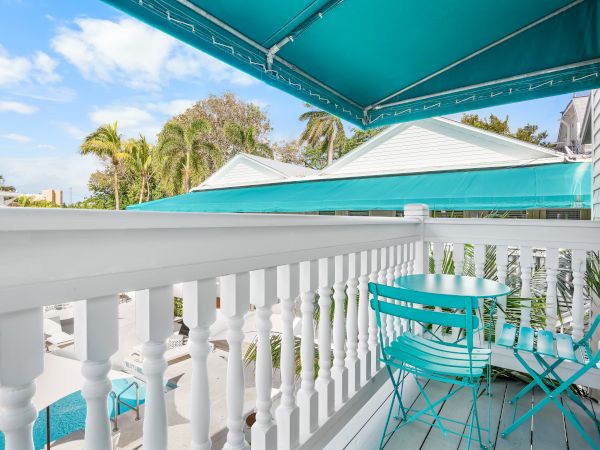 The image shows a balcony with turquoise chairs and table, a white railing, awning, and a view of rooftops and palm trees on a sunny day.