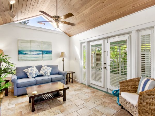 A cozy living room with a blue sofa, coffee table, wicker chair, and ceiling fan. Glass doors lead outside. Art decorates the wall.