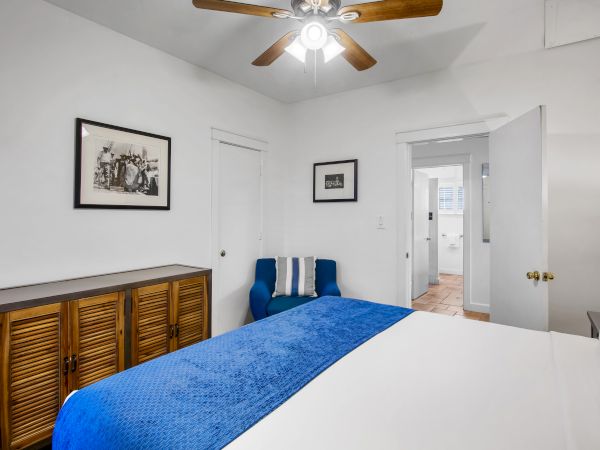 A bedroom with a blue bedspread, wooden cabinet, artwork on the walls, and a ceiling fan. There's also a blue chair near the corner.