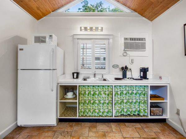 A small kitchenette with a fridge, microwave, sink, and shelves with floral curtains. Coffee makers and a window are above the sink.