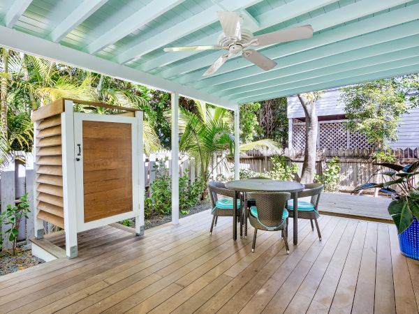 The image shows a covered outdoor patio with a ceiling fan, table and chairs, wooden flooring, and surrounding greenery in a backyard setting.