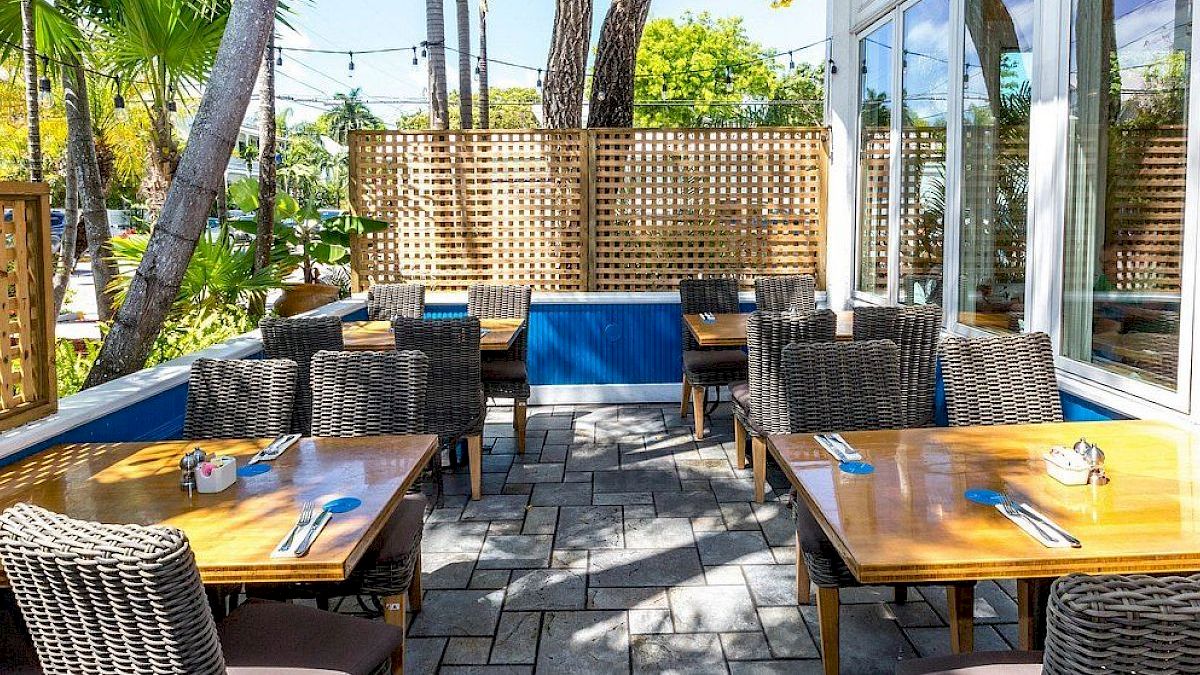 Outdoor patio dining area with wooden tables, wicker chairs, and lush greenery. Sunny and inviting atmosphere.
