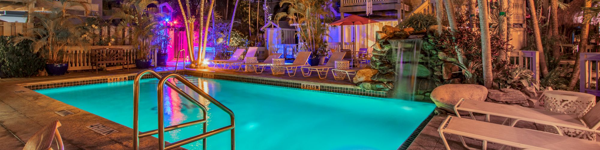 A tropical pool area at night, surrounded by palm trees and lounge chairs, illuminated by colorful lights.