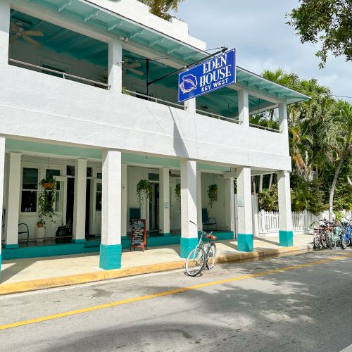The image shows a white building with teal accents and a blue "Hun House" sign, surrounded by palm trees and parked bicycles on the street.