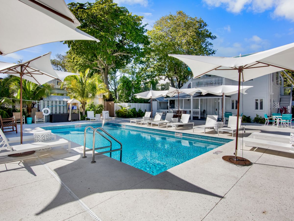 A sunny hotel poolside scene with clear blue water, lounge chairs, white umbrellas, and palm trees, surrounded by light-colored buildings.