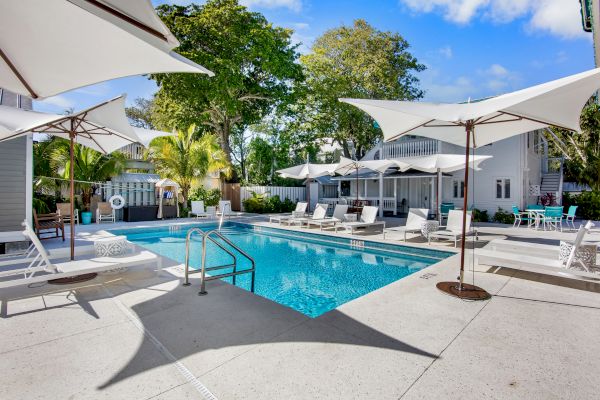 The image shows a serene pool area with lounge chairs, umbrellas, and surrounding trees under a clear blue sky.