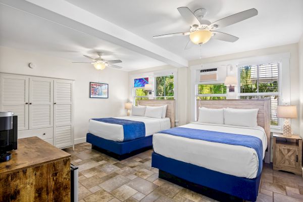 The image shows a bedroom with two double beds, blue bed runners, ceiling fans, lamps, a dresser, and windows with shutters.