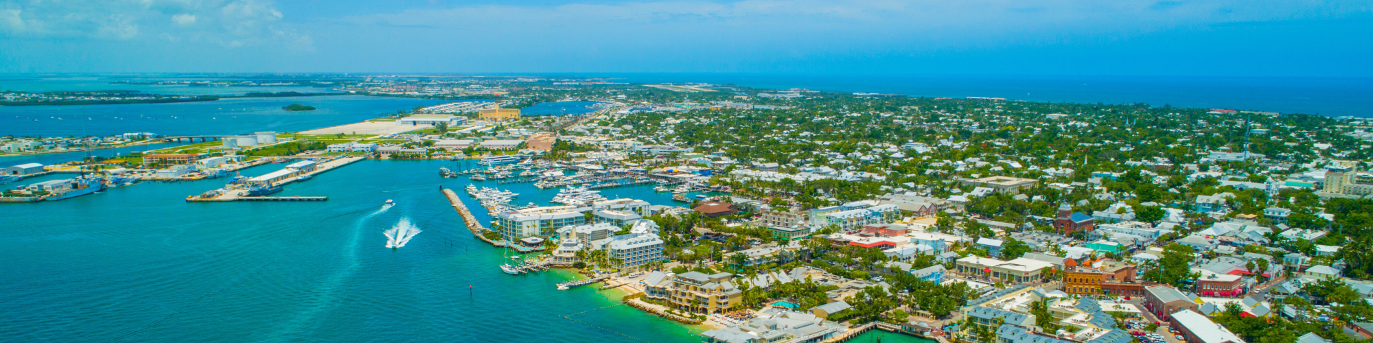 Aerial view of a coastal city with turquoise waters, docks, boats, and urban buildings under a clear, blue sky.