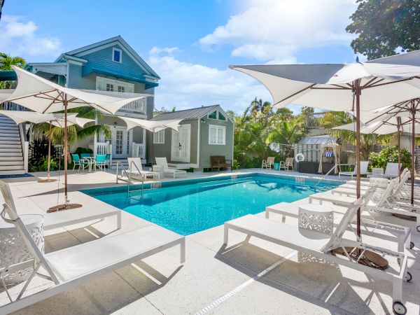 The image features a pool area with white lounge chairs and large umbrellas, surrounded by a light-colored deck and a charming blue house.