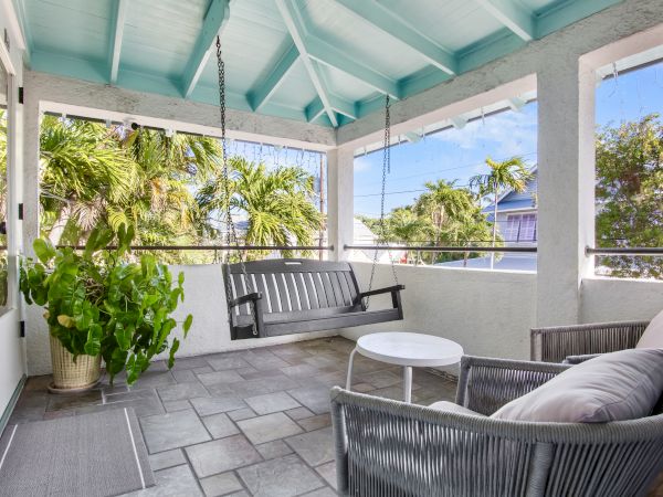 A cozy porch with a swing, wicker chairs, a small table, and lush plants. The ceiling is painted light blue, offering a tropical feel.