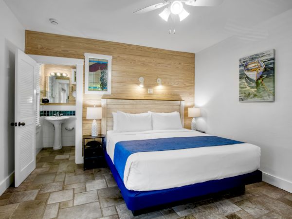 A modern bedroom with a large bed, blue bedspread, wall art, ceiling fan, and an open door leading to a bathroom with a sink and mirror.