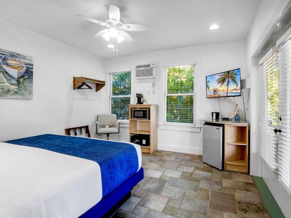 A bright room with a bed, TV, mini fridge, microwave, and decor. Features tiles, fan, and window shades. Tropical image on TV screen.