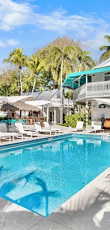 A sunny hotel-style courtyard with a rectangular blue pool, white poolside chairs, sun umbrellas, and pastel two-story buildings with teal awnings.