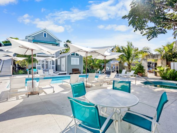 Outdoor pool area with teal chairs, white umbrellas, and a bright house surrounded by trees and plants under a clear blue sky.